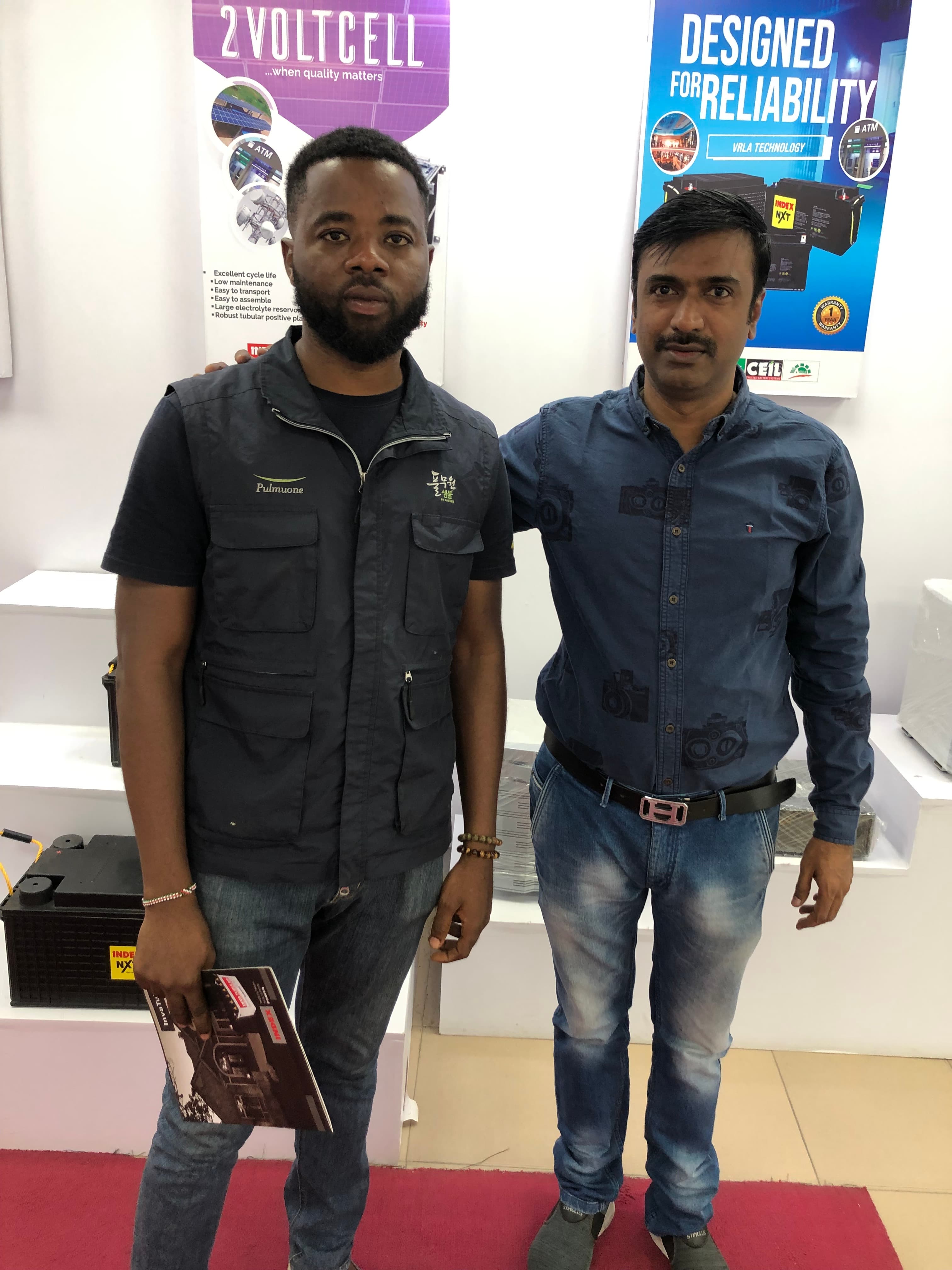 Two men standing in a battery showroom with promotional posters, one holding a product brochure.